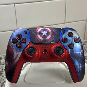 May include: A custom red, blue, and black video game controller featuring Captain America's shield and other Marvel-themed graphics. The controller has red buttons and joysticks, with a clear stand.