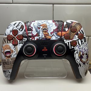 May include: Customized black and white PlayStation 5 controller featuring anime-style graphics. The controller has red and brown accents on the buttons and a central image of a character. The design includes the text "WANTED" and "DEAD OR ALIVE".