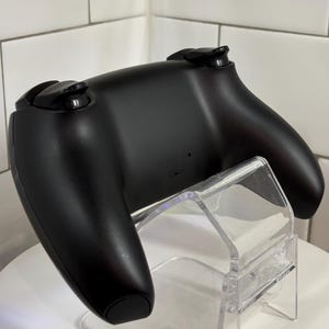 May include: A black video game controller with two analog sticks and several buttons. The controller is resting on a clear acrylic stand, showcasing its ergonomic design. The background is a white tiled wall.