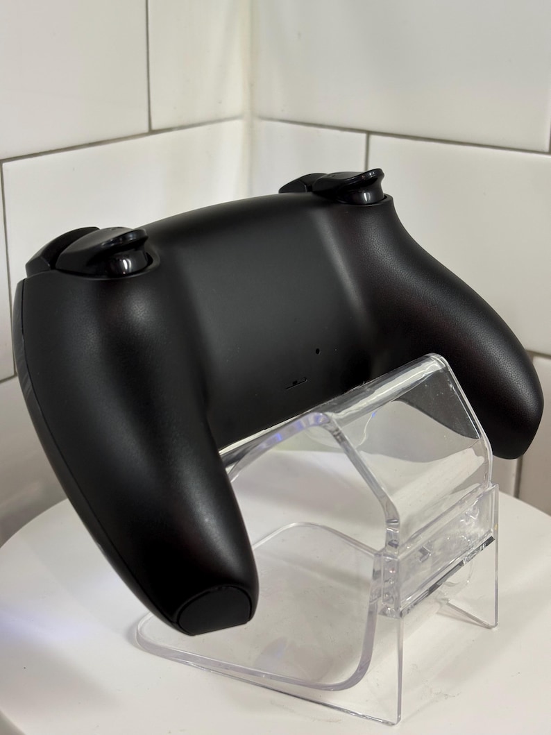 May include: A black video game controller with two analog sticks and several buttons. The controller is resting on a clear acrylic stand, showcasing its ergonomic design. The background is a white tiled wall.
