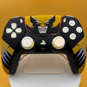 May include: A black and yellow wireless game controller featuring a graphic of a character with a mask and sharp teeth on the touchpad. The controller includes white analog sticks, a directional pad, and action buttons with unique symbols.