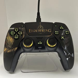 May include: Black gaming controller with the text "ELDEN RING" in gold. The controller has gold accents around the joysticks and green illuminated buttons. The controller is connected to a black cable.