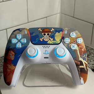 Könnte beinhalten: Ein weiß-blauer PlayStation 5-Controller mit einem individuellen Design mit Charakteren und Bildern aus der Anime-Serie One Piece. Der Controller hat blau beleuchtete Tasten und ein Totenkopf-Logo.