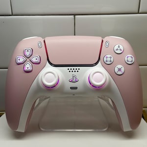 Pode incluir: Um comando PlayStation 5 personalizado em rosa blush e branco. O comando apresenta um touchpad, botões e punhos rosa, com detalhes brancos. Os botões direcionais e os manípulos analógicos têm um brilho roxo. O logótipo PlayStation está centrado.