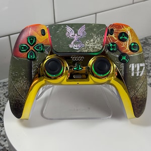 Puede incluir: Mando de videojuegos personalizado con un diseño dorado y verde oliva. El mando presenta el logotipo de UNSC y el número 117. Los botones y joysticks tienen detalles verdes.