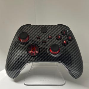 Peut inclure: Une manette de jeu vidéo noire et grise texturée en fibre de carbone avec des boutons d'accentuation rouges. La manette possède un pavé directionnel central et quatre boutons d'action étiquetés X, Y, A et B. La manette est exposée sur un support transparent.