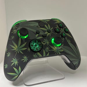 Op de afbeelding: Een zwarte Xbox-controller met een groen marihuanabladpatroon. De controller heeft groene LED-lampjes op de knoppen en de D-pad.