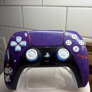 Puede incluir: Un mando de PlayStation 5 morado y negro con un diseño de Kuromi. El mando presenta botones blancos, detalles en azul y la palabra "Kuromi" en el lado derecho. Se muestra sobre un soporte transparente.