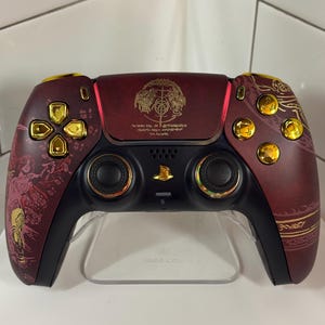 Puede incluir: Un mando de videojuegos personalizado en burdeos y negro con detalles dorados. El mando presenta diseños y símbolos intrincados, con botones dorados y un emblema central. El mando se muestra sobre un soporte transparente.