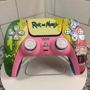May include: A custom pink and green PlayStation 5 controller with "Rick and Morty" text and character art. The controller has white joysticks and a clear stand.