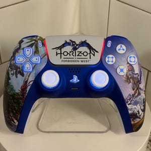 Pode incluir: Um comando PlayStation 5 azul e branco com um design "Horizon Forbidden West". O comando apresenta um motivo de palmeira e personagem nas laterais, com o logótipo do jogo no centro. Os botões e joysticks têm um brilho azul.