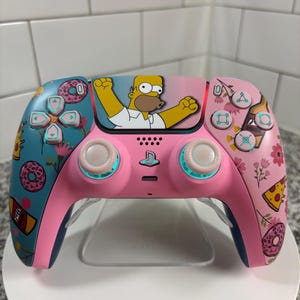 Könnte beinhalten: Ein rosa-blauer Videospiel-Controller mit einem Cartoon-Bild von Homer Simpson. Der Controller hat ein Blumen- und Lebensmittel-Design mit Donuts und Pizza. Die Knöpfe sind türkis und rosa.