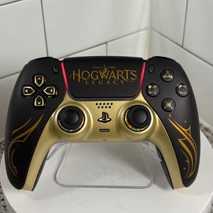 May include: Black and gold PlayStation controller featuring the text "Hogwarts Legacy." The controller has gold detailing on the buttons and around the joysticks. It is displayed on a clear stand.