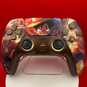 May include: A wireless game controller featuring a custom wood-grain finish and vibrant anime-style graphics of a character with a straw hat. The controller includes black analog sticks, gold-colored directional buttons, and a central touchpad display.