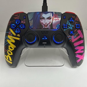 May include: A black PlayStation 5 DualSense controller with a custom design featuring a cartoon character with pink and blue hair. The controller has a blue light around the touchpad and the buttons. The text "Boom!" and "Tinx Here" are written on the controller.