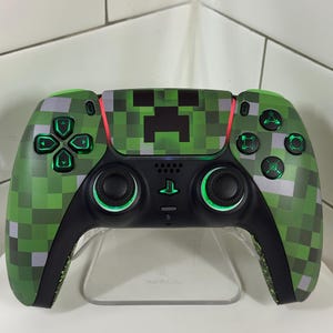 以下が含まれることがあります： 緑色のピクセル化されたデザインのカスタムビデオゲームコントローラー。中央に黒いクリーパーの顔があります。コントローラーには緑色の照明付きボタンとジョイスティックがあり、黒いベースが付いています。透明なスタンドに表示されています。
