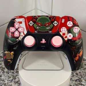May include: A black and red video game controller featuring Teenage Mutant Ninja Turtles graphics. The controller has pink-glowing thumbsticks and a white base. The design includes images of the turtles and various symbols.