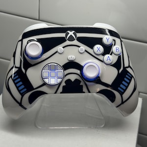 May include: White and black Xbox controller with a Stormtrooper design. The controller has blue accents and a glossy finish. The design features the iconic helmet and details of the character.