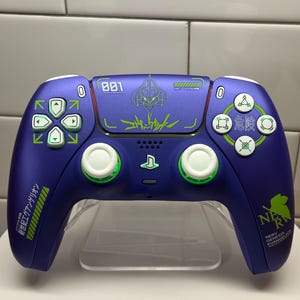 Könnte beinhalten: Ein benutzerdefinierter lila PlayStation 5-Controller mit grünen und weißen Akzenten. Der Controller verfügt über japanischen Text und Grafiken, einschließlich des Textes "NERV" und "EVANGELION". Die Tasten sind weiß und grün, mit einem Steuerkreuz und Aktionstasten.