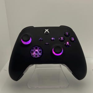 May include: A black Xbox controller with a purple light around the buttons and a textured grip. The controller is on a clear stand.