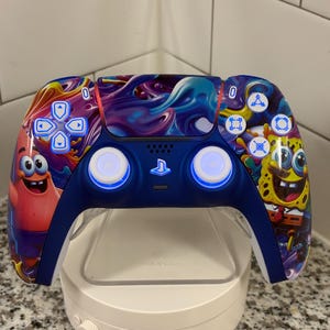 May include: A custom blue and white video game controller with cartoon characters Patrick Star and SpongeBob SquarePants. The controller has glowing white buttons and is on a white charging stand.