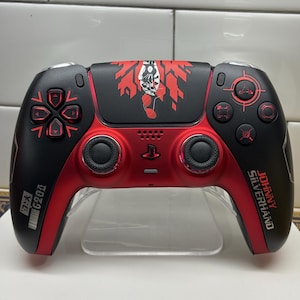 Op de afbeelding: Een aangepaste PlayStation 5-controller in zwart en rood. De controller heeft rode knoppen, een D-pad en accenten. De bovenkant heeft een rood en wit grafisch ontwerp. De tekst "Johnny Silverhand" is zichtbaar.