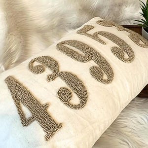 May include: Cream rectangular cushion embroidered with the number "4366" in textured, light brown thread. The cushion rests on white faux fur, with a textured grey cushion in the background.