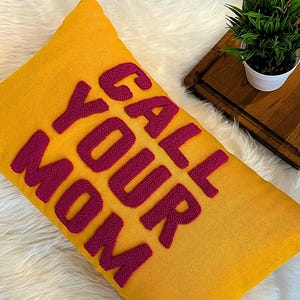 May include: A yellow throw pillow with the phrase "Call Your Mom" embroidered in pink.