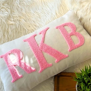 Könnte beinhalten: Ein weißes Kissen mit den Buchstaben "RKB" in rosa gestickt. Die Buchstaben bestehen aus flauschigem, geschlungenem Garn.