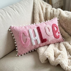 May include: A pink gingham cushion embroidered with the name "CHLOE" in shades of pink and white. The cushion has a pom-pom trim and is placed on a beige sofa, with a fluffy blanket draped over it. The cushion is approximately 40 cm long.