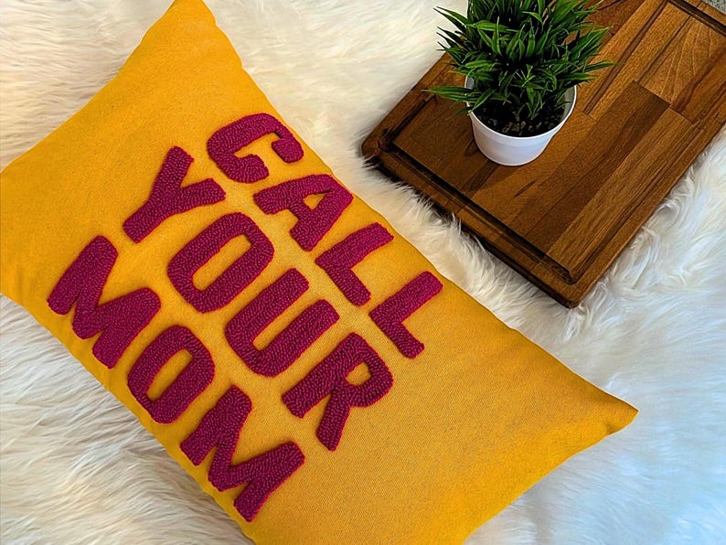 May include: A yellow throw pillow with the phrase "Call Your Mom" embroidered in pink.