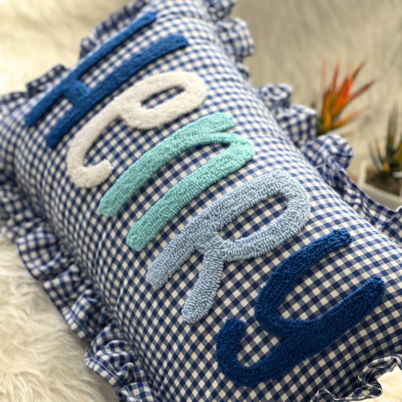 May include: A blue and white gingham pillow with the word "Happy" embroidered in textured, varying shades of blue and white. The pillow has a ruffled edge.