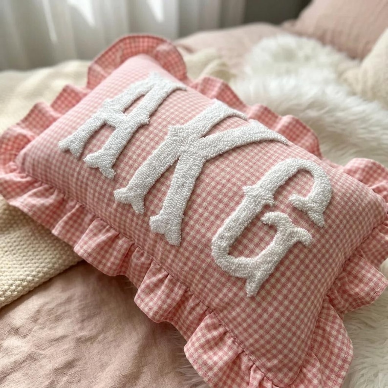 May include: A rectangular throw pillow featuring a light pink and white gingham pattern with a ruffled border. The center displays the large, white, textured, embroidered letters A, K, and G in a decorative, serif-style font across the fabric.