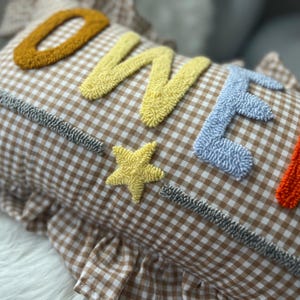 May include: A decorative pillow with the word "ONE" in yellow, blue, and orange textured letters. The pillow has a brown and white gingham pattern and a yellow star. The pillow has a ruffled edge.
