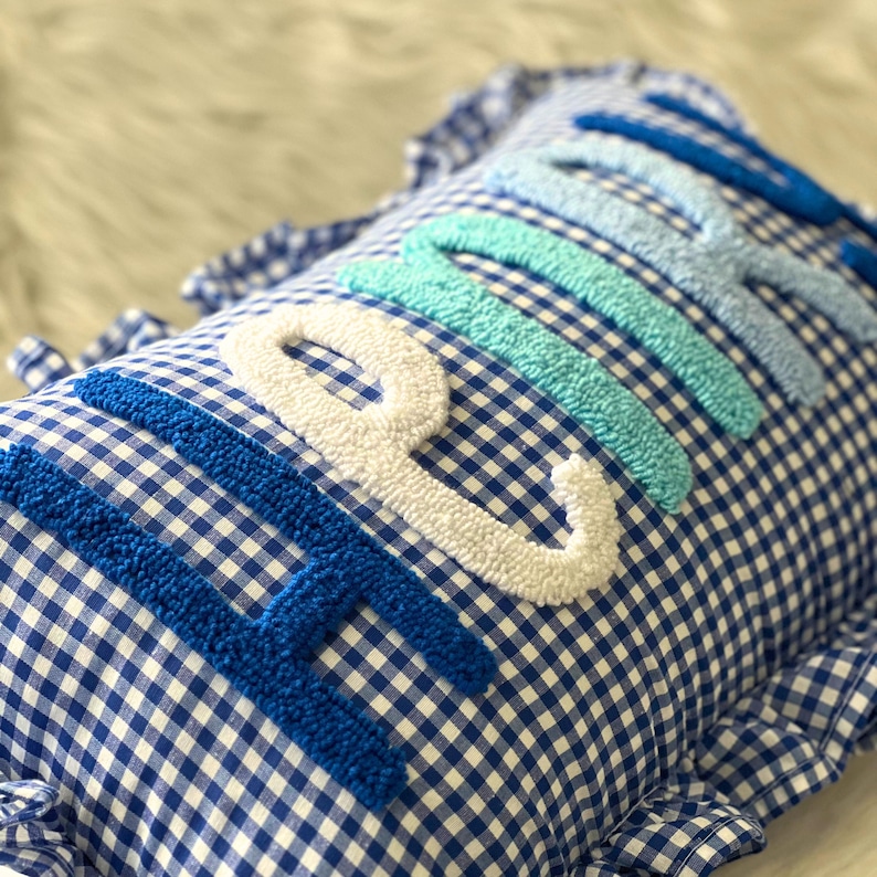 May include: Blue and white gingham pillow with the word "BEACH" embroidered in varying shades of blue and white. The pillow has a ruffled edge and is designed for decorative use.