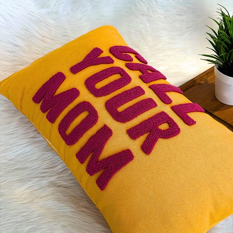 May include: A yellow throw pillow with the phrase "Grab Your Mom" embroidered in pink.