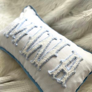 May include: White rectangular pillow with the embroidered word "MAMA" in light blue and white. The pillow has a light blue border. The letters are in a decorative font.
