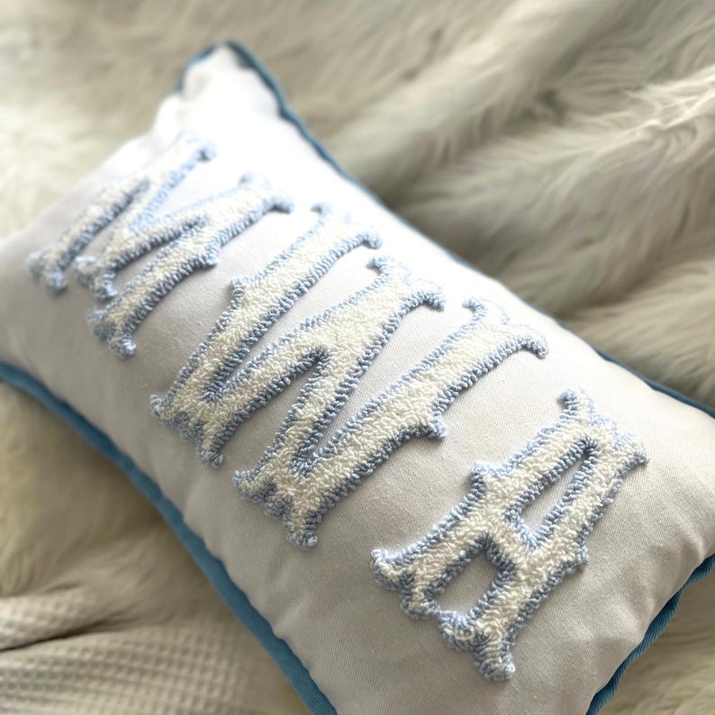 May include: White rectangular pillow with the embroidered word "MAMA" in light blue and white. The pillow has a light blue border. The letters are in a decorative font.