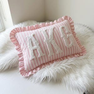 May include: A rectangular throw pillow featuring a light pink and white vertical striped pattern. The pillow has a ruffled border and displays the letters AKG in large, white, textured, tufted fabric appliqu&eacute;s centered on the front surface.