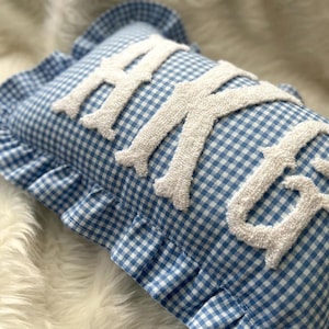 May include: A rectangular throw pillow featuring a blue and white gingham check pattern with a ruffled edge. The surface displays the word HUGS in large, white, textured, tufted letters that are raised from the fabric of the pillow cover.