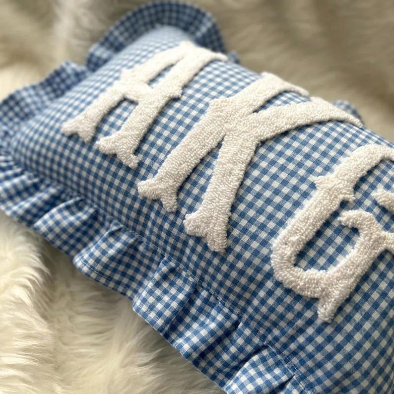 May include: A rectangular throw pillow featuring a blue and white gingham check pattern with a ruffled edge. The surface displays the word HUGS in large, white, textured, tufted letters that are raised from the fabric of the pillow cover.