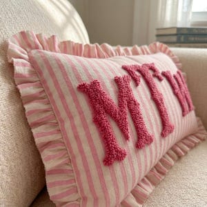May include: A rectangular throw pillow featuring a light pink and white striped pattern with a ruffled border. The front of the pillow displays the letters MTW in raised, textured, dark pink tufted embroidery across the center of the fabric.