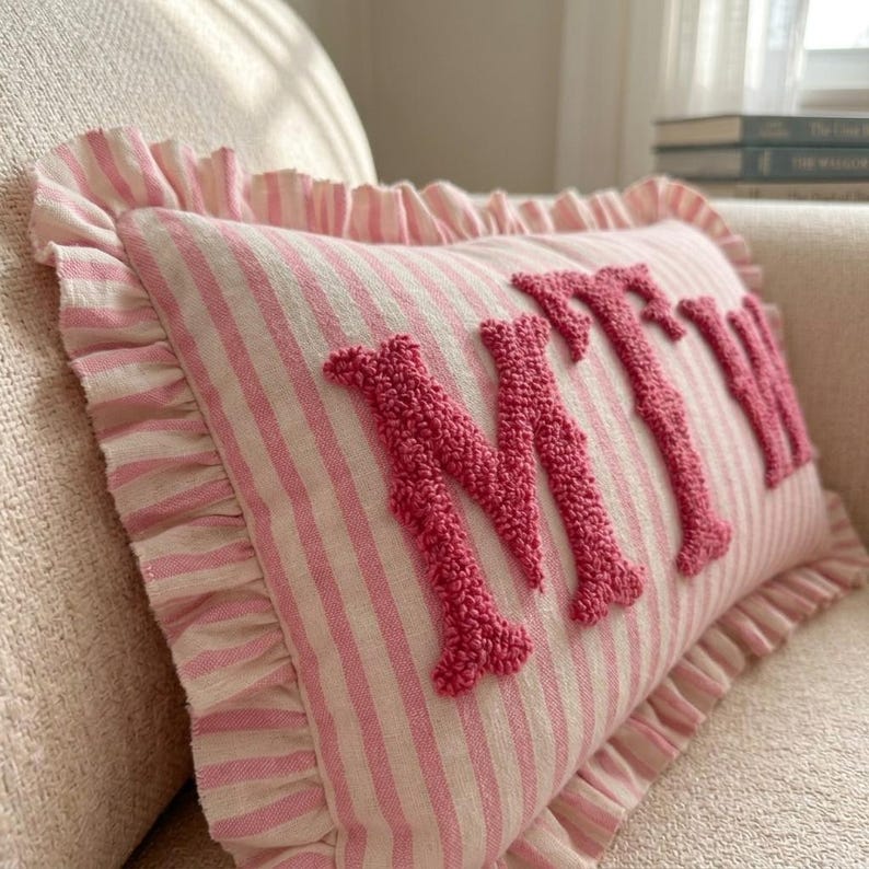 May include: A rectangular throw pillow featuring a light pink and white striped pattern with a ruffled border. The front of the pillow displays the letters MTW in raised, textured, dark pink tufted embroidery across the center of the fabric.