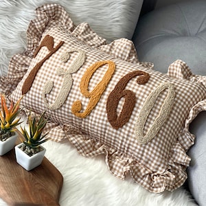 May include: A rectangular decorative pillow with a brown and white gingham pattern. The pillow features the embroidered word "1896" in various shades of brown and tan. The pillow has a ruffled edge and is placed on a white, fluffy surface.