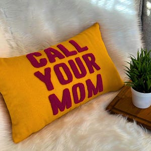 May include: A yellow throw pillow with the words "CALL YOUR MOM" embroidered in pink.