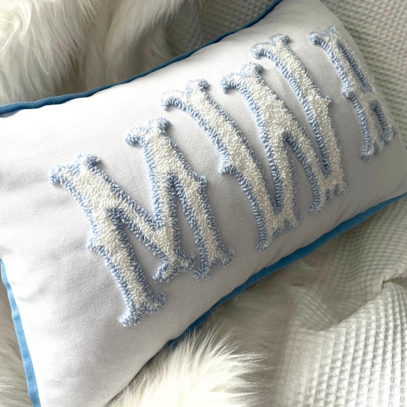 May include: White rectangular pillow with the word "MAMA" embroidered in white and light blue. The pillow has a light blue border and is placed on a white, textured surface.
