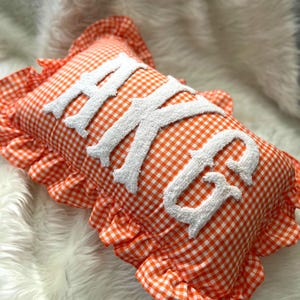 Può includere: Cuscino rettangolare a quadretti arancioni e bianchi con le lettere bianche ricamate "AKG". Il cuscino ha un bordo a balze e poggia su una superficie bianca e soffice.