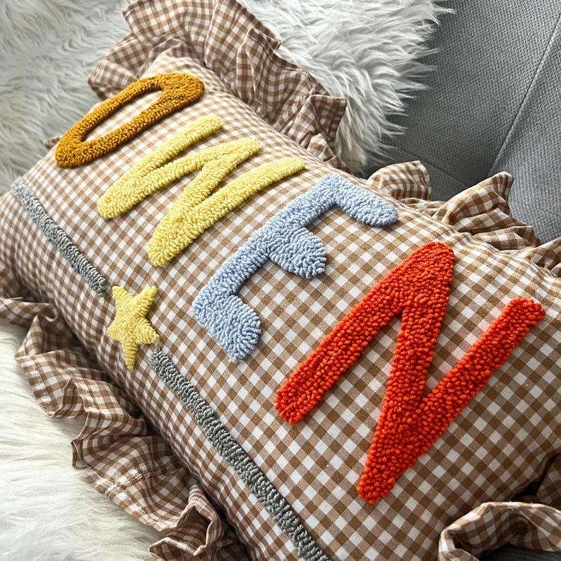 May include: A brown and white gingham pillow with the word 'ONE' embroidered in colorful yarn. The letters are in a rainbow color scheme, with the 'O' in orange, the 'N' in yellow, and the 'E' in blue. The pillow has a ruffled edge and a small star in the corner.