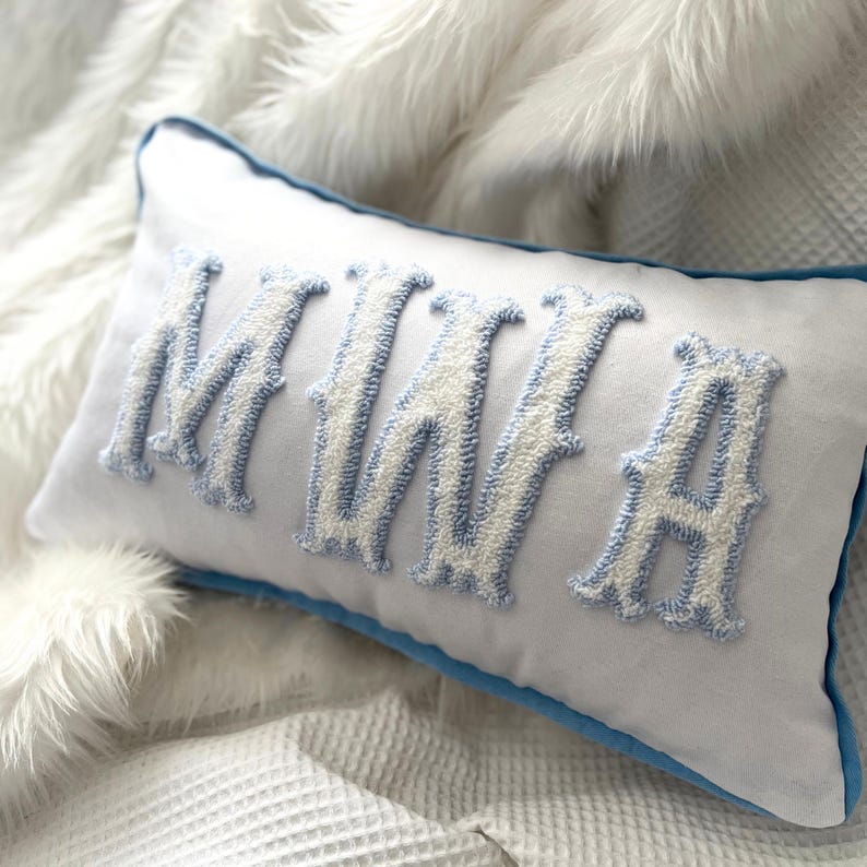 May include: A rectangular white pillow with the name "MINA" embroidered in white and light blue. The letters are in a decorative font and have a textured appearance. The pillow has a light blue border and is placed on a white, textured surface.