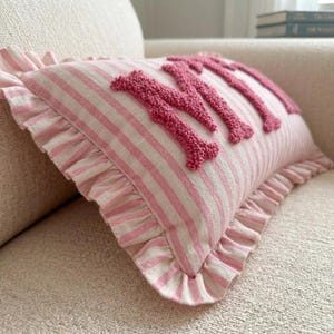 May include: A rectangular throw pillow featuring a light pink and white striped fabric with a ruffled edge. The front of the pillow displays the word "Mimi" embroidered in a raised, textured, dark pink yarn with a looped pile finish.